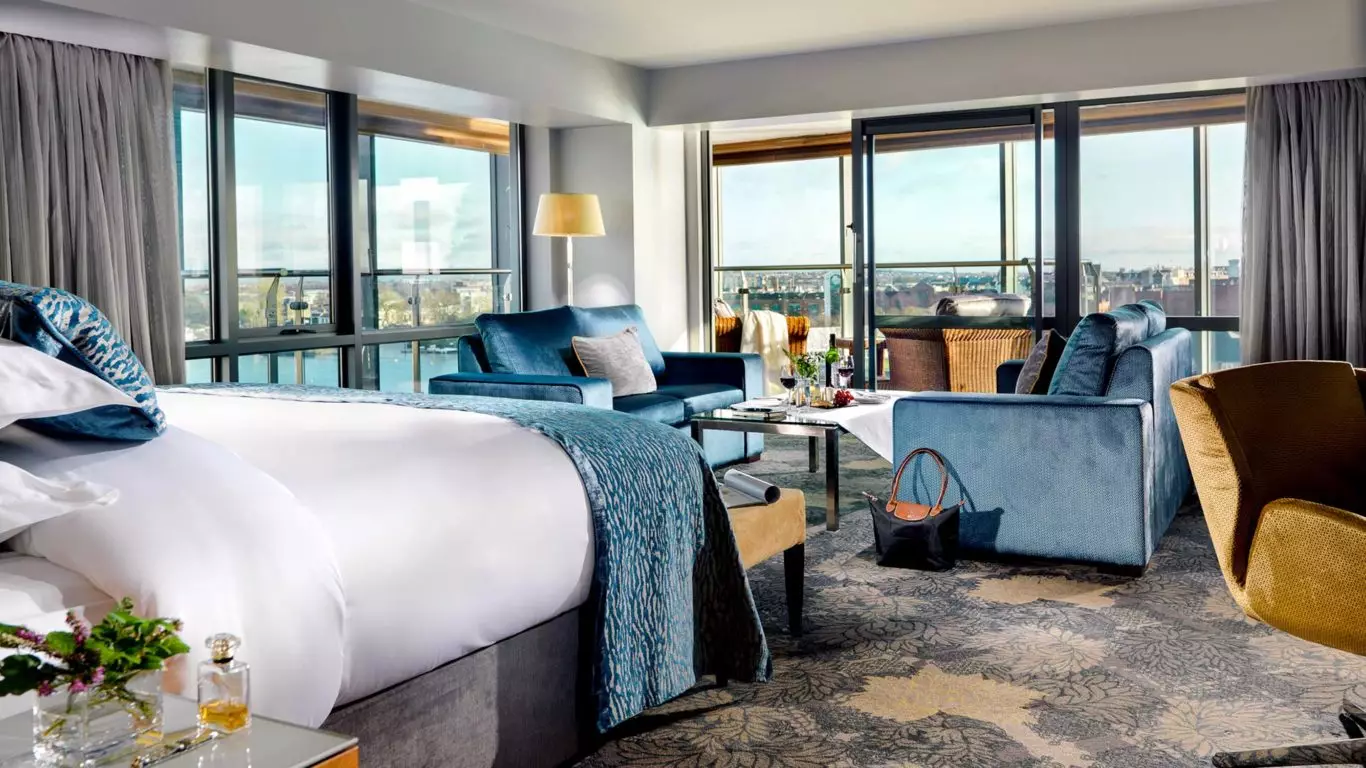 Spacious hotel room with a large bed, seating area, floor-to-ceiling windows, and a city view. Blue and beige decor, with a purse placed on the floor near a coffee table.