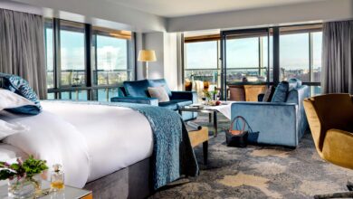 Spacious hotel room with a large bed, seating area, floor-to-ceiling windows, and a city view. Blue and beige decor, with a purse placed on the floor near a coffee table.