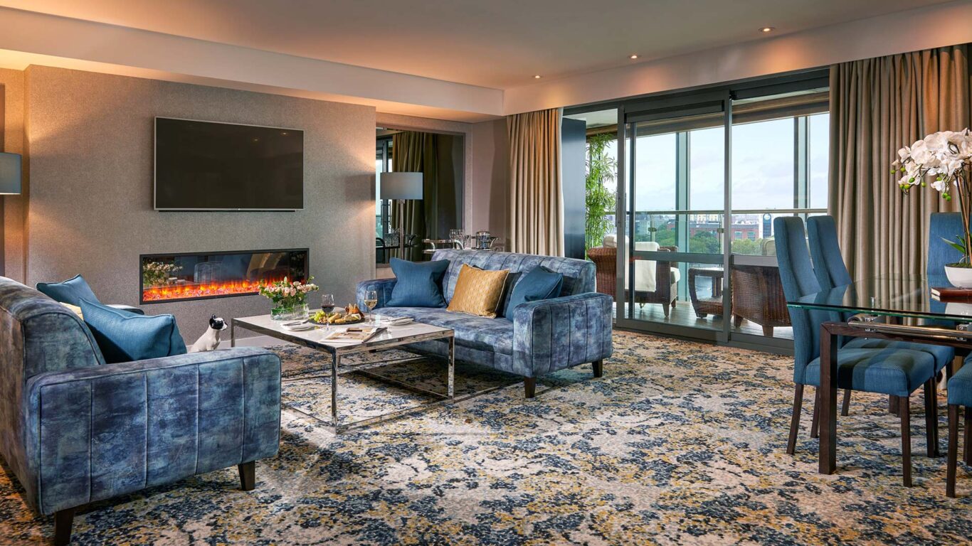 Modern hotel suite with blue sofas, a wall-mounted TV above an electric fireplace, a glass dining table, large windows with curtains, and a balcony view.
