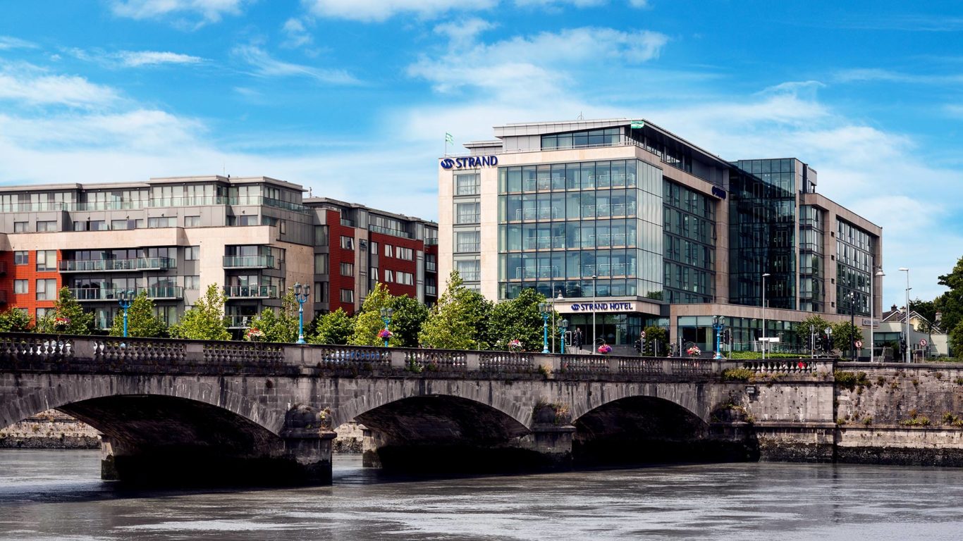Modern glass-fronted hotel next to a bridge over a river, with adjacent apartment buildings, trees, and a partly cloudy blue sky.