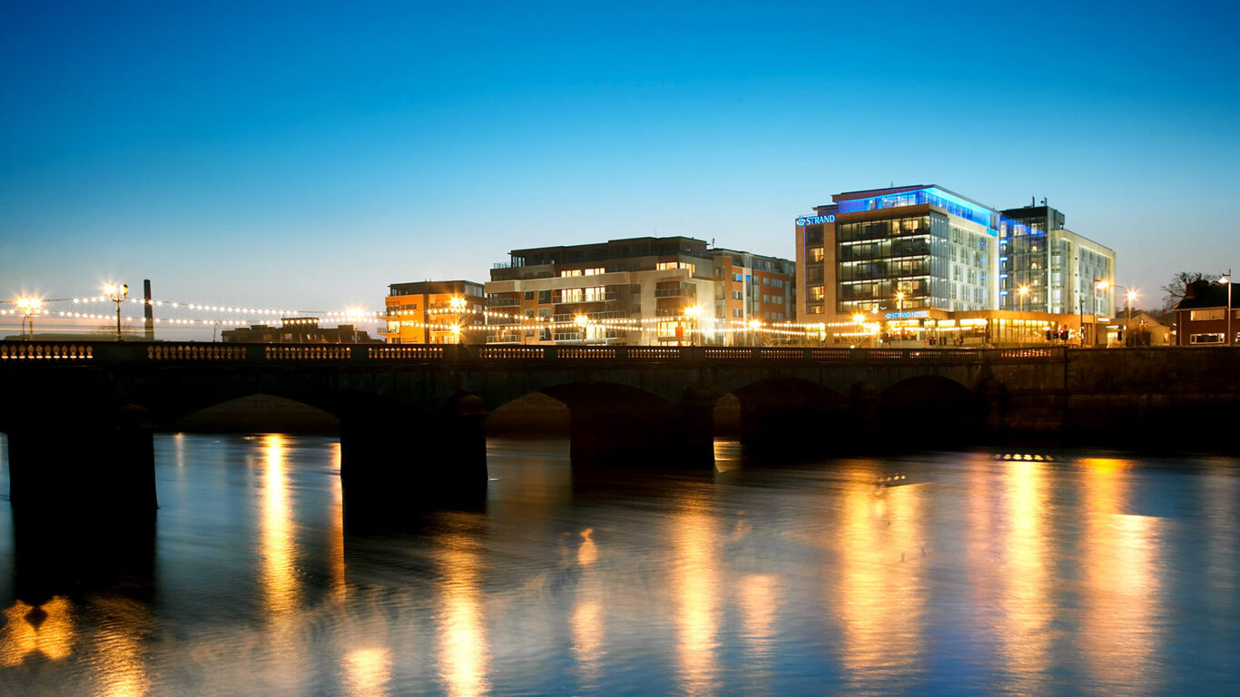 A modern riverside hotel and adjacent buildings are illuminated at dusk, with lights reflecting on the calm water below a bridge.