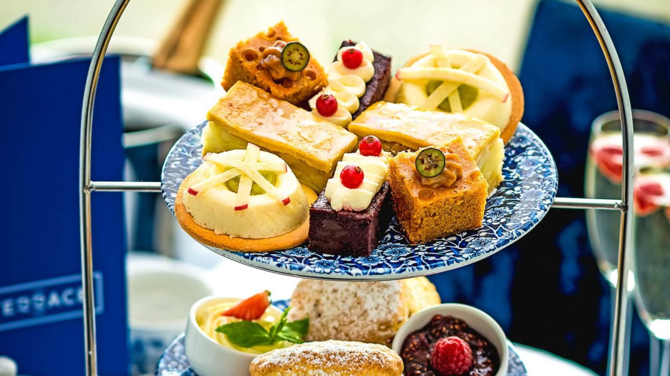 A two-tiered tray displays assorted pastries, cakes, and scones with fruit garnishes, cream, and jam on a blue-patterned plate.