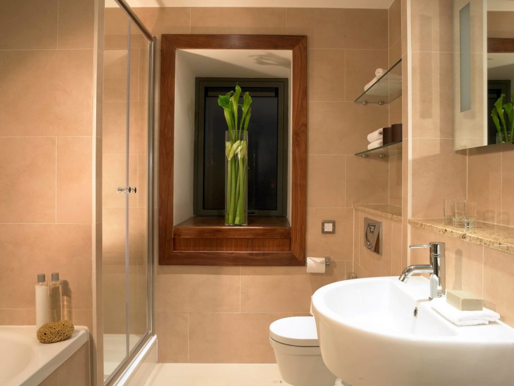 Modern bathroom with beige tiles, glass shower, white sink and toilet, and a vase with tall green plants on the wooden window ledge.