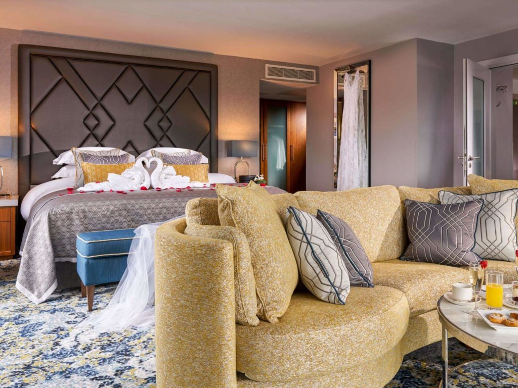 A modern hotel room with a large bed, swan-shaped towels, a yellow sofa, decorative pillows, a small table with drinks, and patterned carpet.