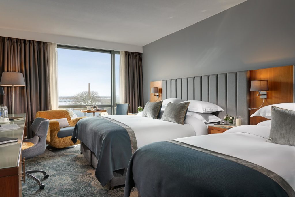 Modern hotel room with two double beds, a desk, armchair, large window with a view, and neutral-toned decor.