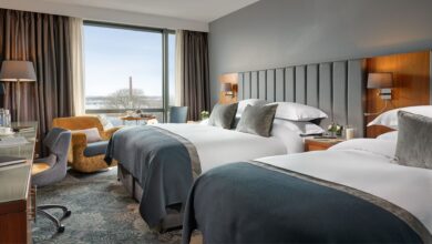 Modern hotel room with two double beds, a desk, chair, lamps, and large window overlooking an outdoor scene with trees and a distant chimney.