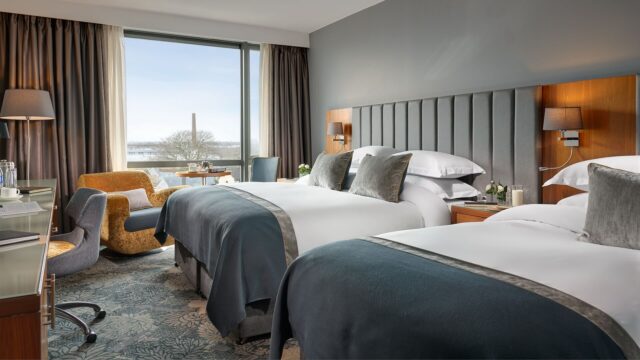 Modern hotel room with two double beds, a desk, chair, lamps, and large window overlooking an outdoor scene with trees and a distant chimney.