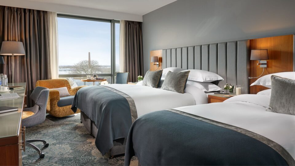 Modern hotel room with two double beds, a desk, chair, lamps, and large window overlooking an outdoor scene with trees and a distant chimney.