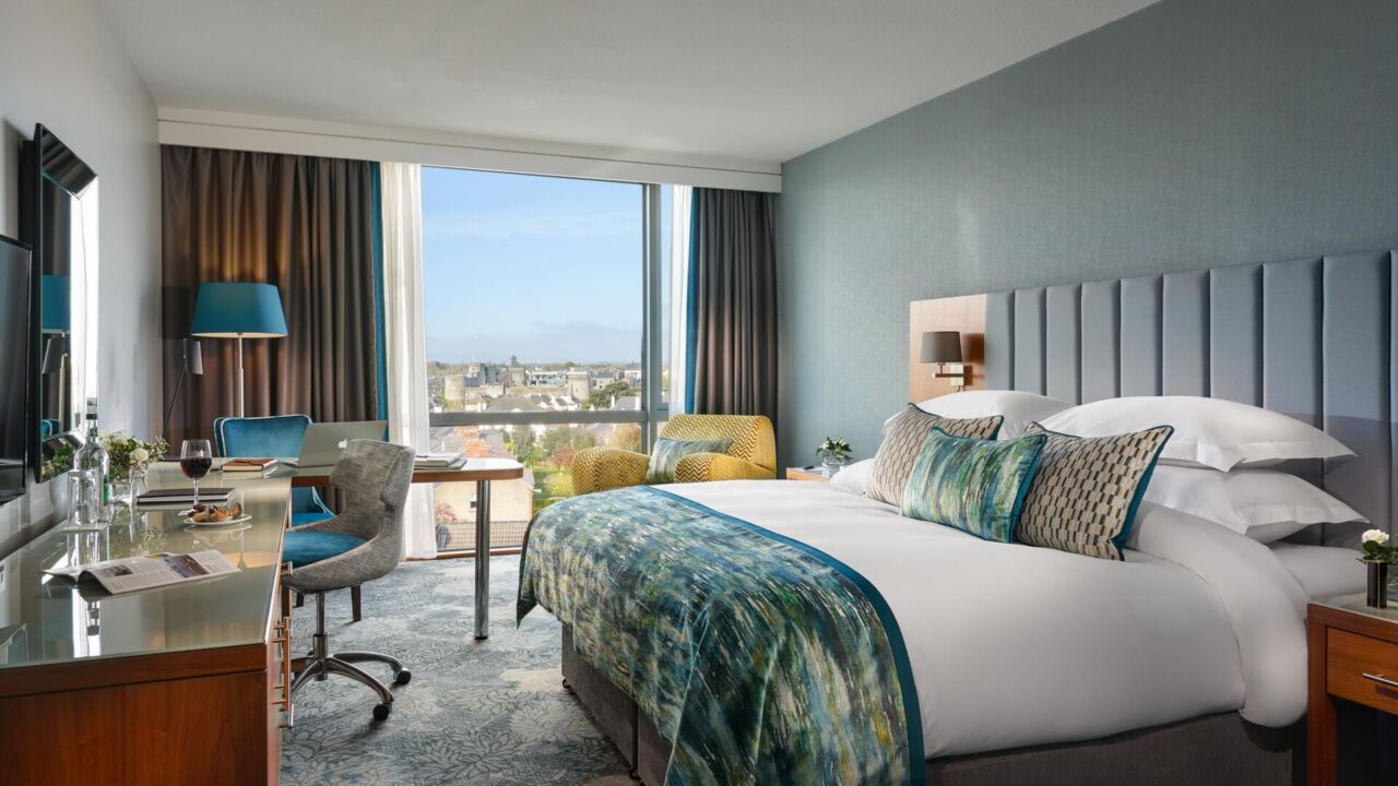 Modern hotel room with a large bed, desk, armchair, floor-to-ceiling window, and city view. Room is well-lit and decorated in blue and grey tones.
