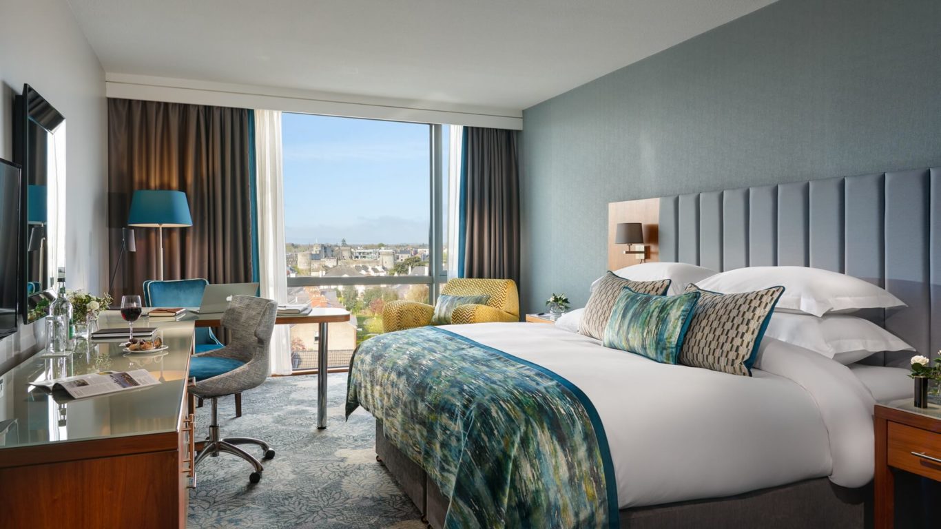 Modern hotel room with a large bed, desk, armchair, and floor-to-ceiling window showing a city view. Room is decorated in blue and grey tones with natural light coming in.