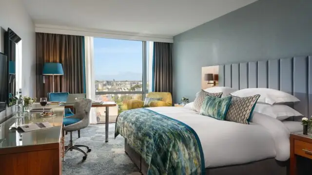 Modern hotel room with a large bed, desk, armchairs, and a window offering a city view. Room features blue and gray tones with natural light entering from the window.