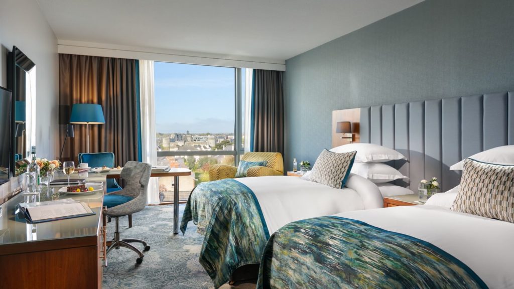 Modern hotel room with two twin beds, a desk, seating area, large window with city view, and contemporary decor in blue and gray tones.