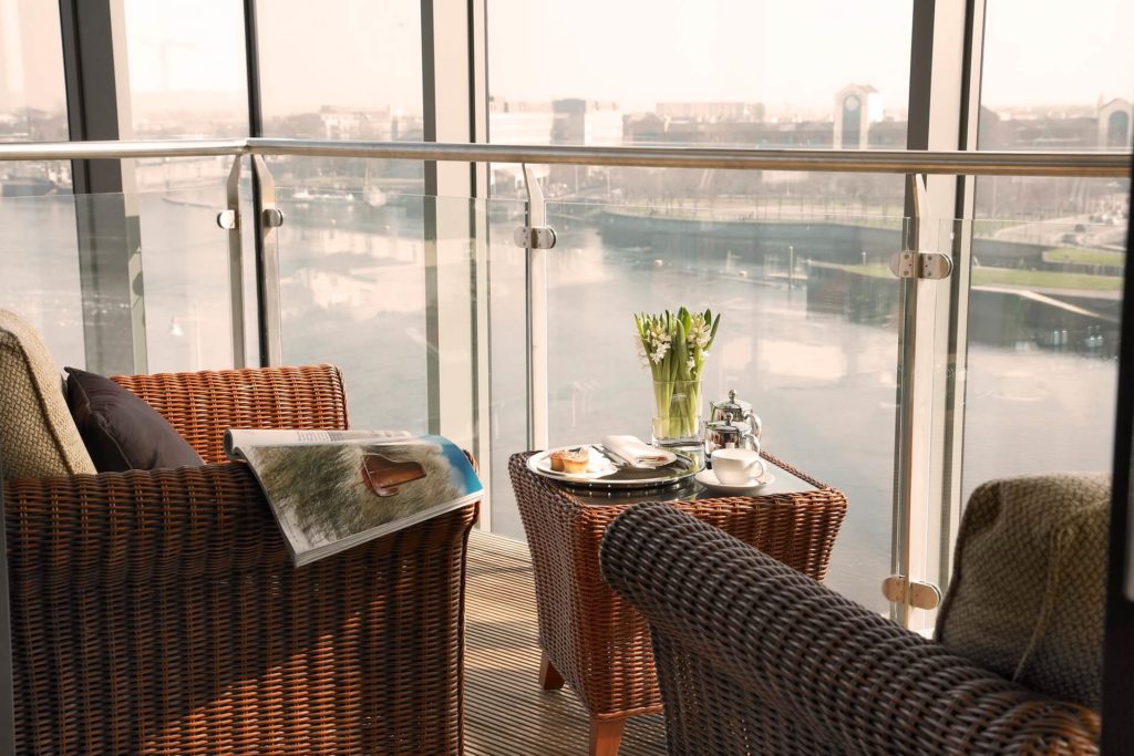 Two wicker chairs and a small table with coffee, flowers, and a magazine are set on a balcony overlooking a river and cityscape through glass panels.