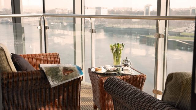 Two wicker chairs and a small table with breakfast and flowers on a balcony overlooking a river and cityscape through glass railings.