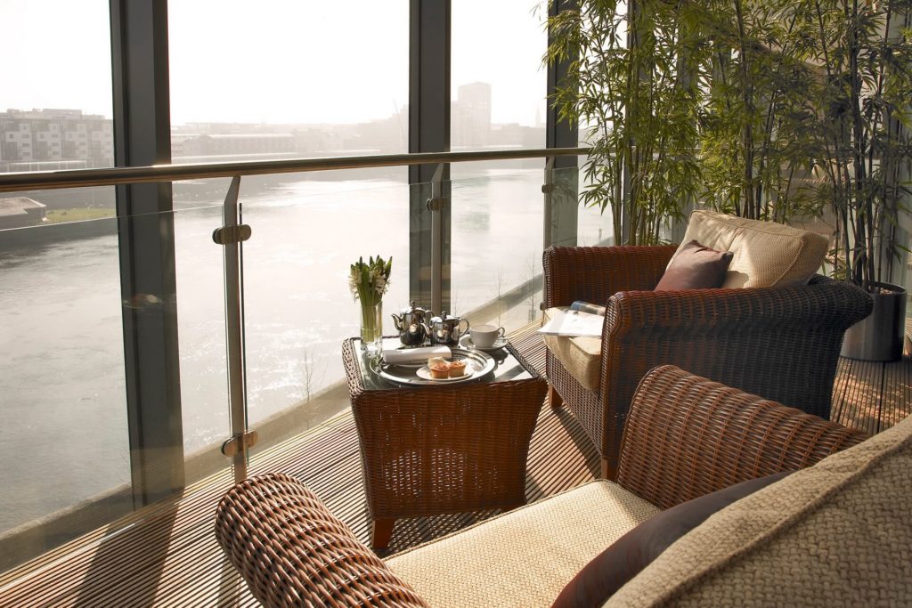 A glass-walled balcony overlooks a river, with wicker chairs, a small table set for tea, and tall potted plants.