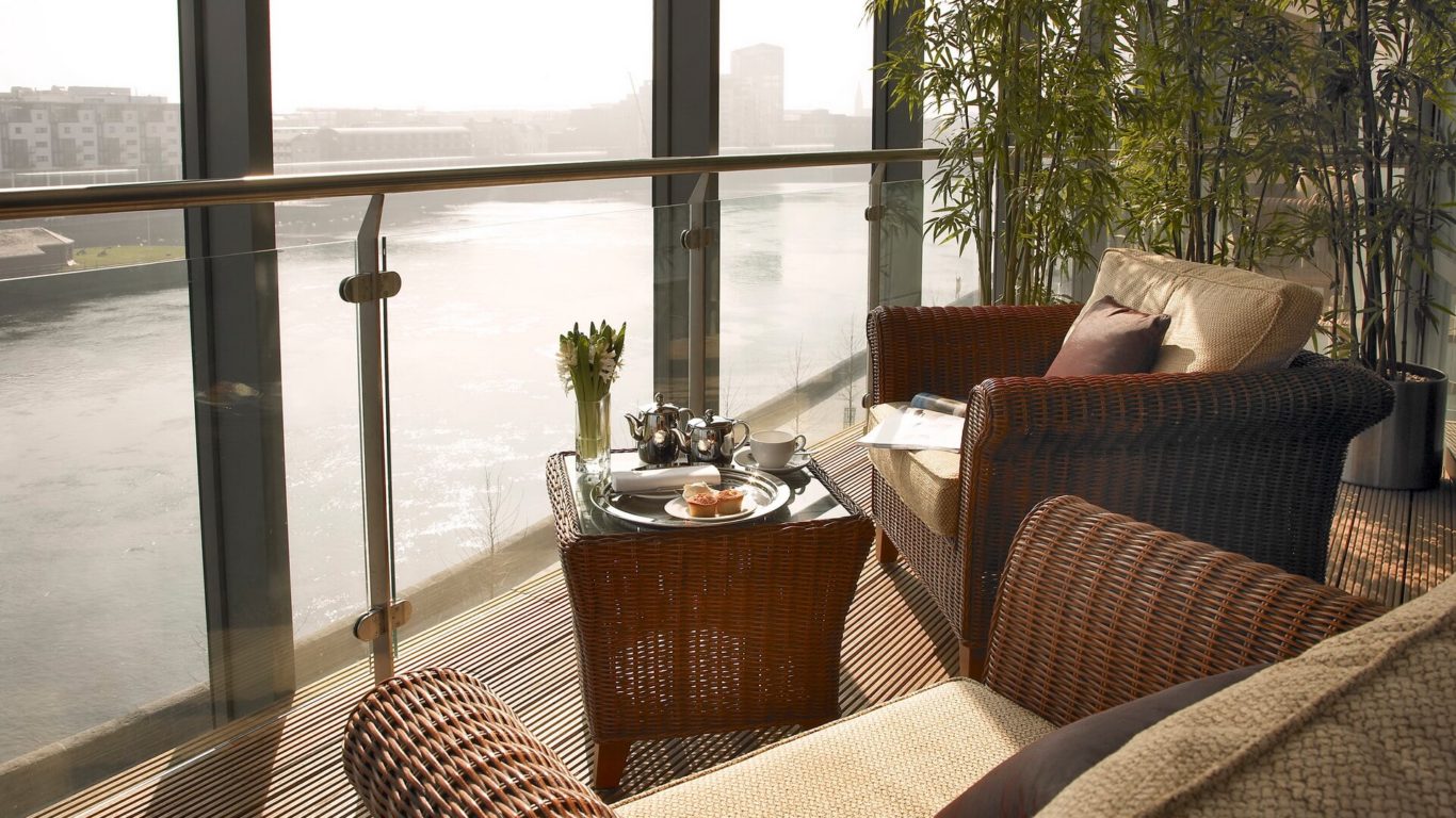 A balcony with wicker chairs, a small table set with tea and pastries, and a view of a river through glass railings.