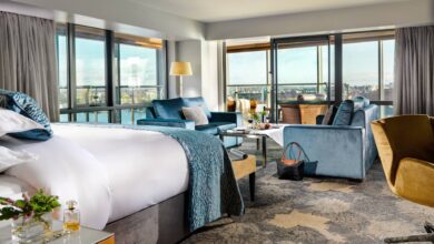 Spacious hotel room with a large bed, blue sofas, armchairs, a coffee table, floor-to-ceiling windows, and a view of the city and river.