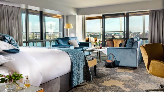 Spacious hotel room with a large bed, blue sofas, armchairs, a coffee table, floor-to-ceiling windows, and a view of the city and river.