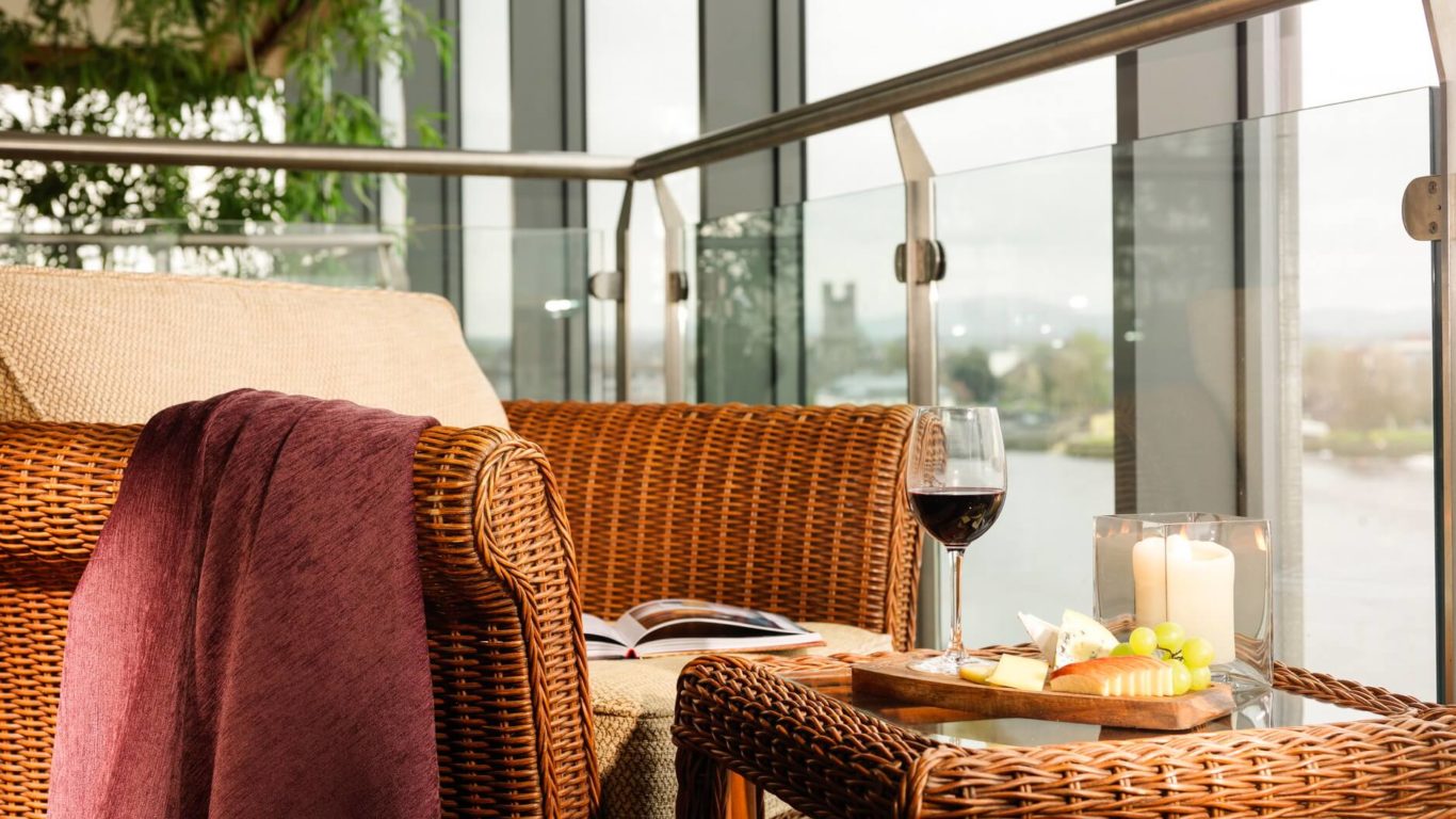 Wicker chair with a purple blanket, an open book, and a glass of red wine next to a plate of cheese and fruit, with candles and a view through large glass windows.
