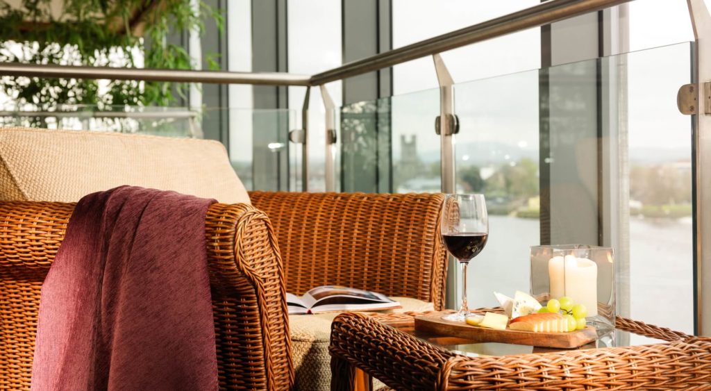 Wicker chair with a maroon throw, an open book, and a tray holding wine, cheese, fruit, and candles on a glass balcony overlooking a river.