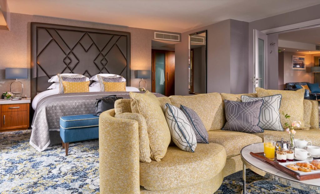 Hotel suite with a large bed, modern headboard, and a yellow curved sofa. A tray with breakfast and drinks sits on a glass coffee table; the room has stylish décor and patterned carpet.