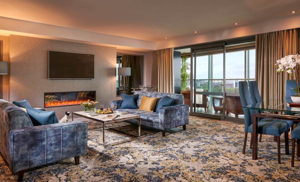 A modern hotel suite with blue sofas, a fireplace under a wall-mounted TV, glass doors opening to a balcony, a dining table, and patterned carpet.