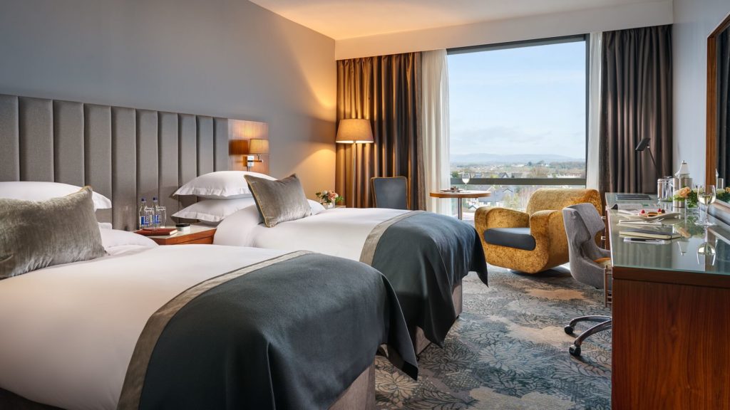 Modern hotel room with two double beds, a desk, an armchair by the window, and a view outside. Bottled water, lamps, and decor are visible on the nightstand and desk.