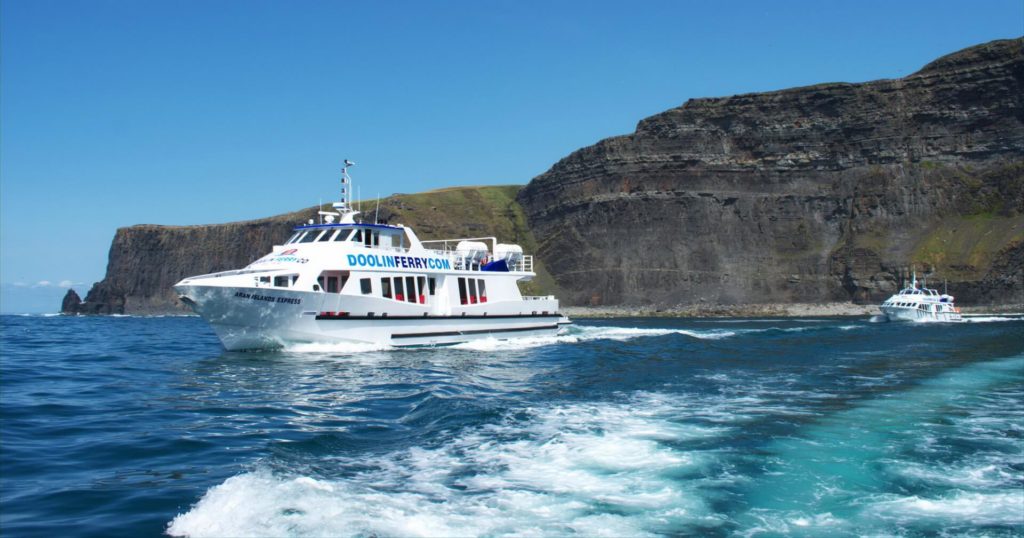 Two white passenger ferries travel on blue water near steep rocky cliffs under a clear sky.