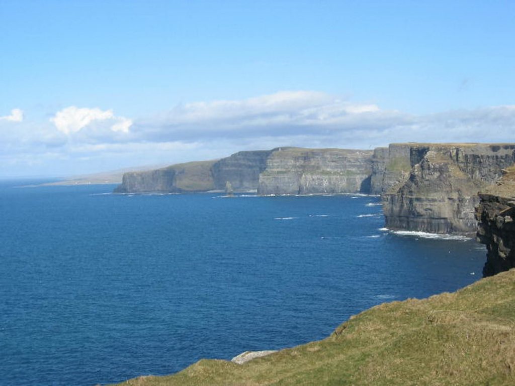Cliffs rise above the blue ocean under a partly cloudy sky, with waves breaking at their base and grassy land in the foreground.