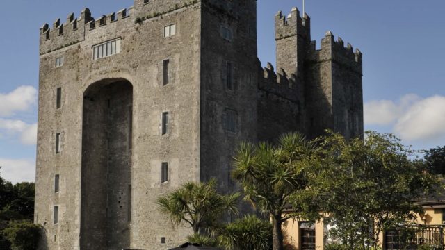 A large stone castle with crenellated towers stands behind a group of people sitting at outdoor tables under a clear blue sky.