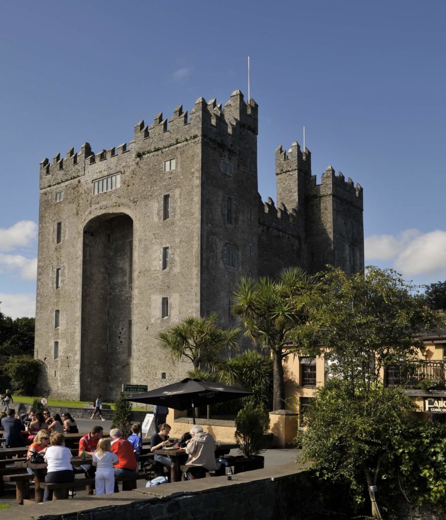 A large stone castle with crenellated towers stands behind a group of people sitting at outdoor tables under a clear blue sky.
