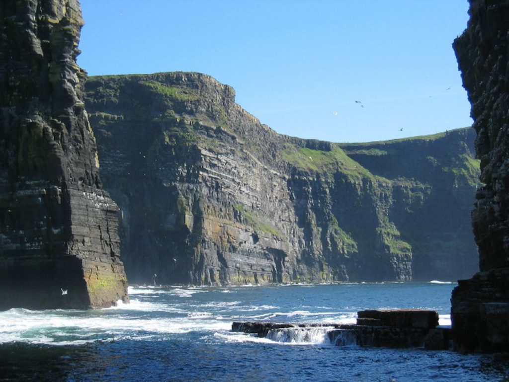 Steep sea cliffs rise above crashing ocean waves under a clear blue sky, with green grass covering the upper edges of the cliffs.