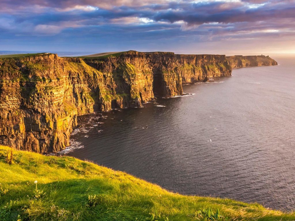 Steep sea cliffs rise above the ocean, with green grass on top and layered rock formations visible, under a partly cloudy sky at sunset.
