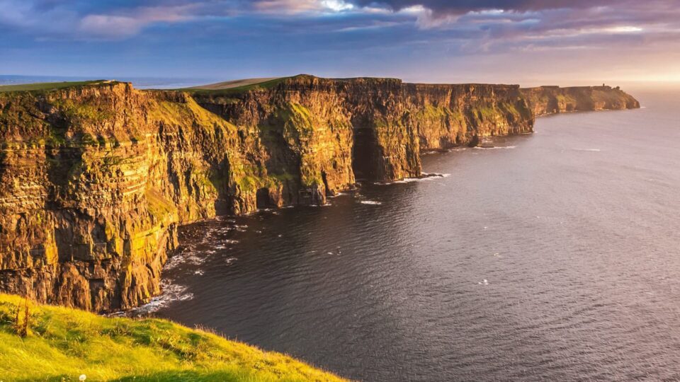 Tall, rugged sea cliffs rise above the Atlantic Ocean, covered in green grass and illuminated by sunlight under a partly cloudy sky.