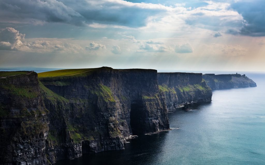 Sheer cliffs rise above the ocean under a partly cloudy sky, with sunlight breaking through and illuminating the green-topped rock faces.