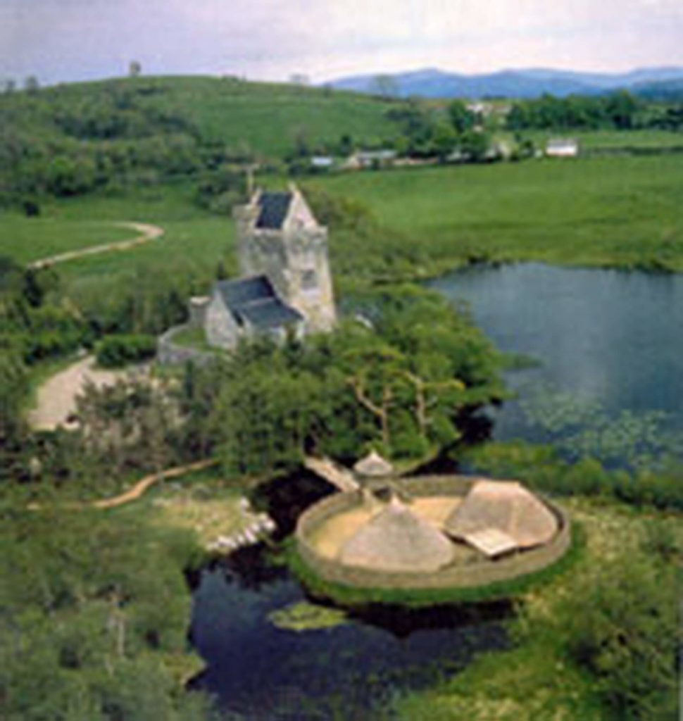 Aerial view of a stone castle and round thatched huts surrounded by water and green fields, with distant hills in the background.