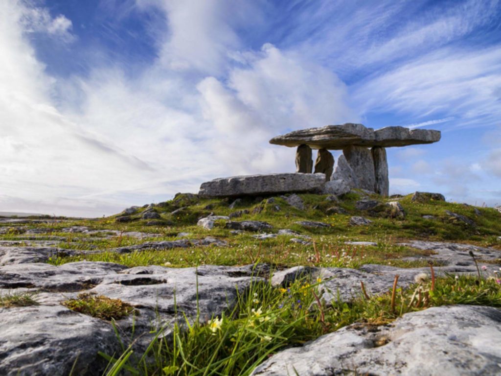 Ancient stone dolmen structure stands on a rocky landscape under a partly cloudy sky with patches of grass in the foreground.