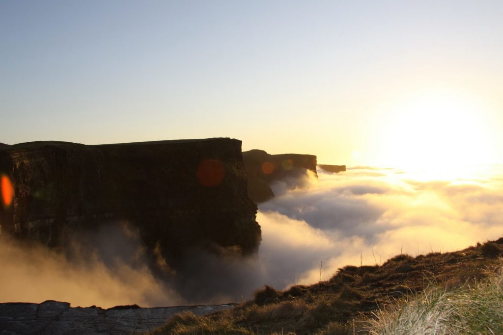 Cliffs rise above a thick layer of clouds at sunrise or sunset, with sunlight illuminating the sky and grass in the foreground.