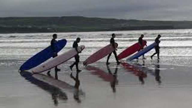 Five people wearing wetsuits walk along a wet, sandy beach carrying surfboards, with the ocean and hills in the background.