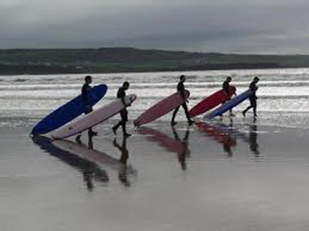 Five people carrying surfboards walk along a wet, reflective beach shoreline under a cloudy sky.