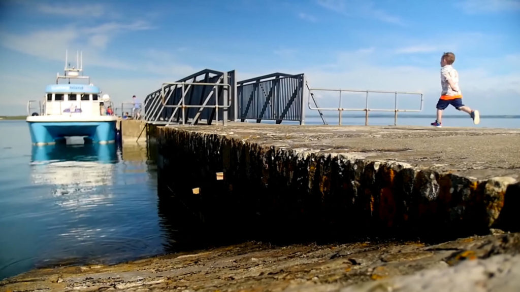A child runs along a concrete pier near a docked boat on a sunny day, with blue sky and water in the background.
