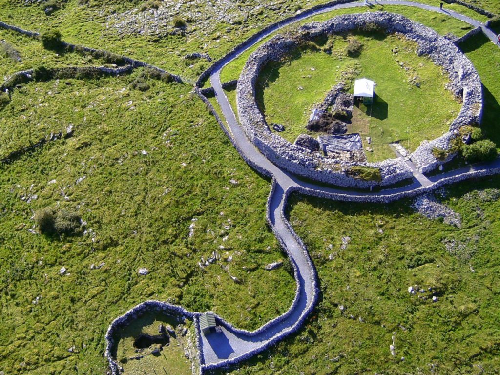 Aerial view of a large circular stone fort with surrounding paths and green landscape, featuring an inner enclosed area and nearby smaller enclosure.