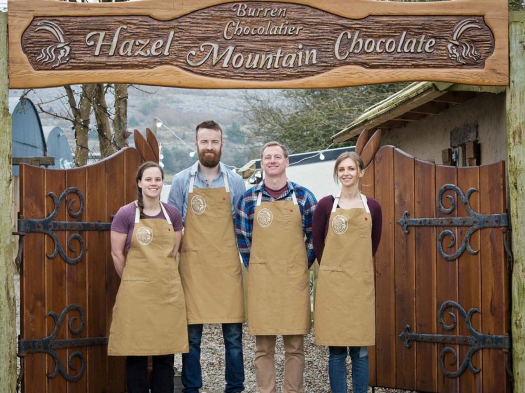 Four people wearing aprons stand in front of a wooden gate and sign that reads 