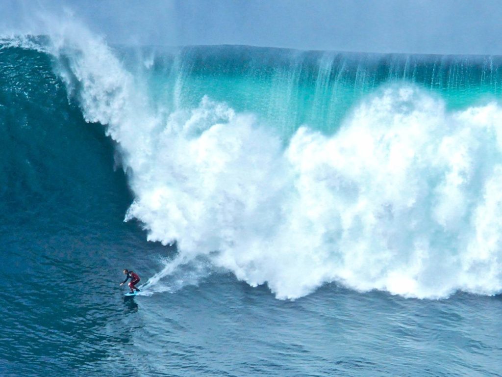 A surfer rides a large, powerful turquoise wave as white spray crashes behind them.
