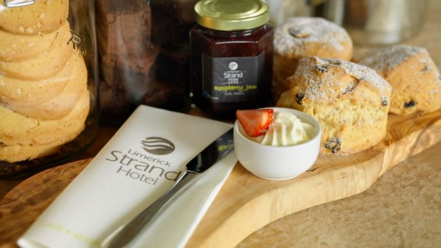 A wooden board with scones dusted with powdered sugar, a bowl of cream topped with a strawberry, a jar of raspberry jam, and a napkin labeled 