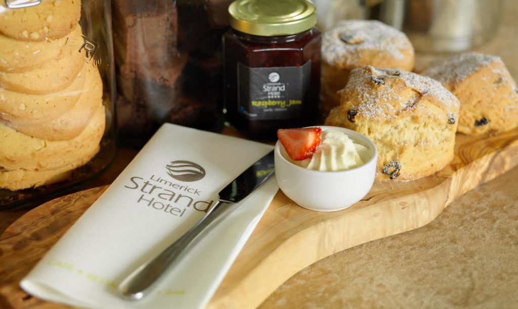 A wooden board with scones, a jar of raspberry jam, a bowl of cream with a strawberry, a knife, and a napkin labeled 
