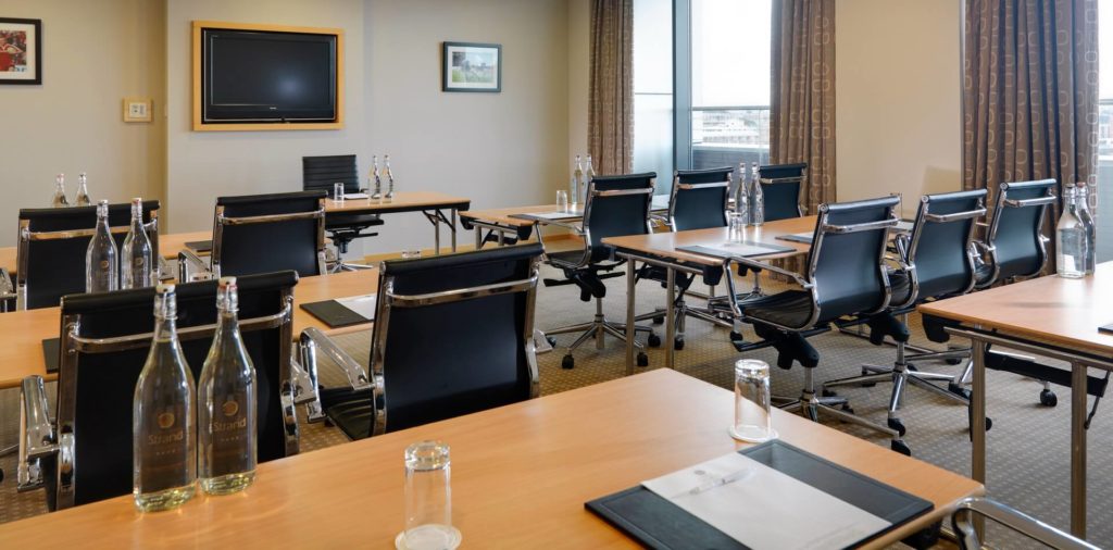 A modern conference room with several tables and black office chairs, each set with bottled water, glasses, notepads, and pens. A wall-mounted TV and framed art are visible.