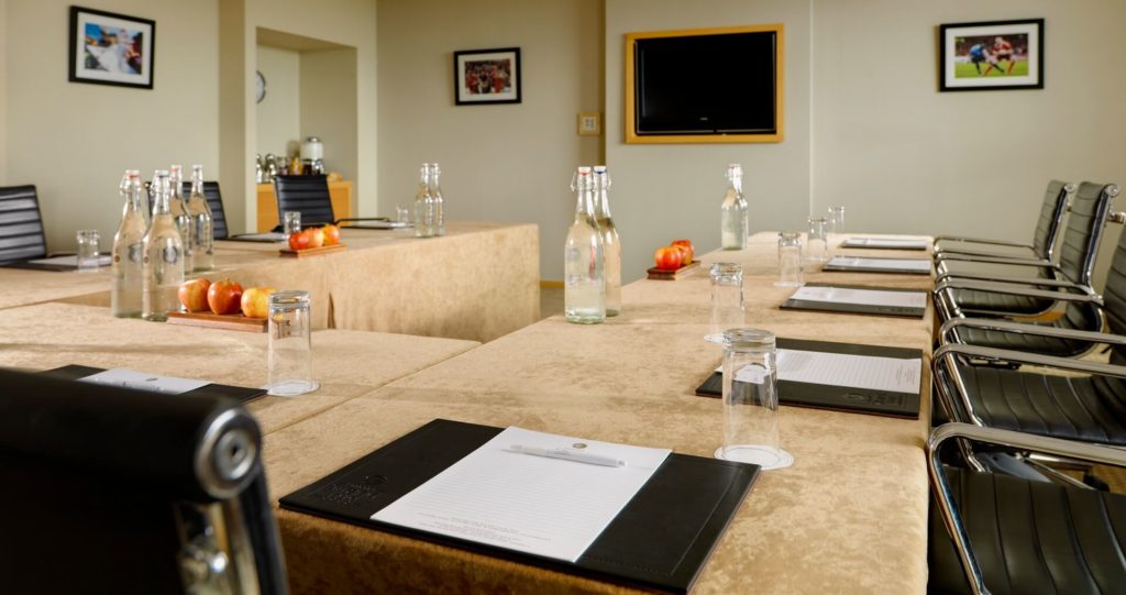 A conference room with tables arranged in a U-shape, set with notepads, pens, water bottles, glasses, and apples. Black chairs are positioned around the tables.