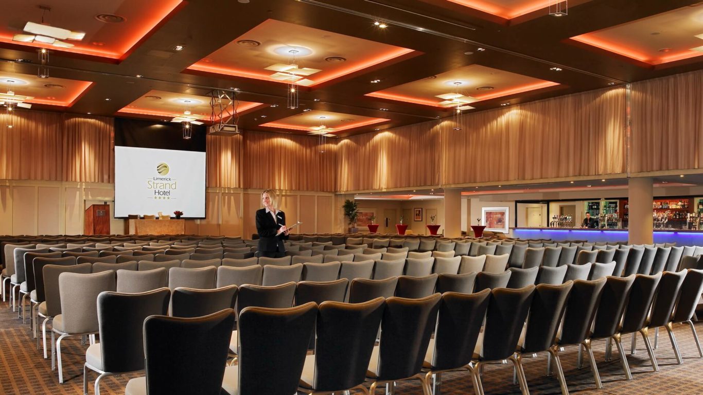A large, empty conference room with rows of chairs facing a stage, a projector screen, and a person standing in the center aisle holding papers.