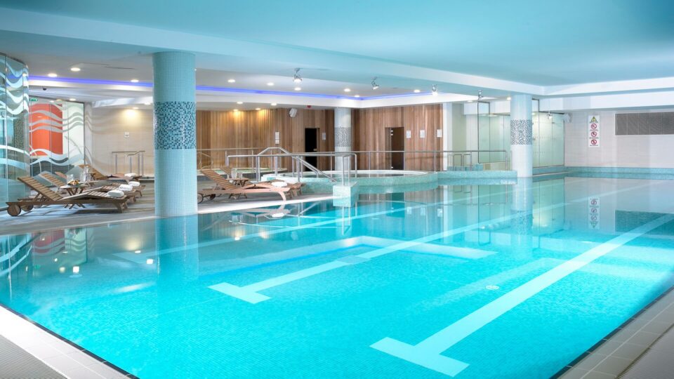 Indoor swimming pool area with lounge chairs, a hot tub, and modern decor; bright lighting reflects off the clear blue water.